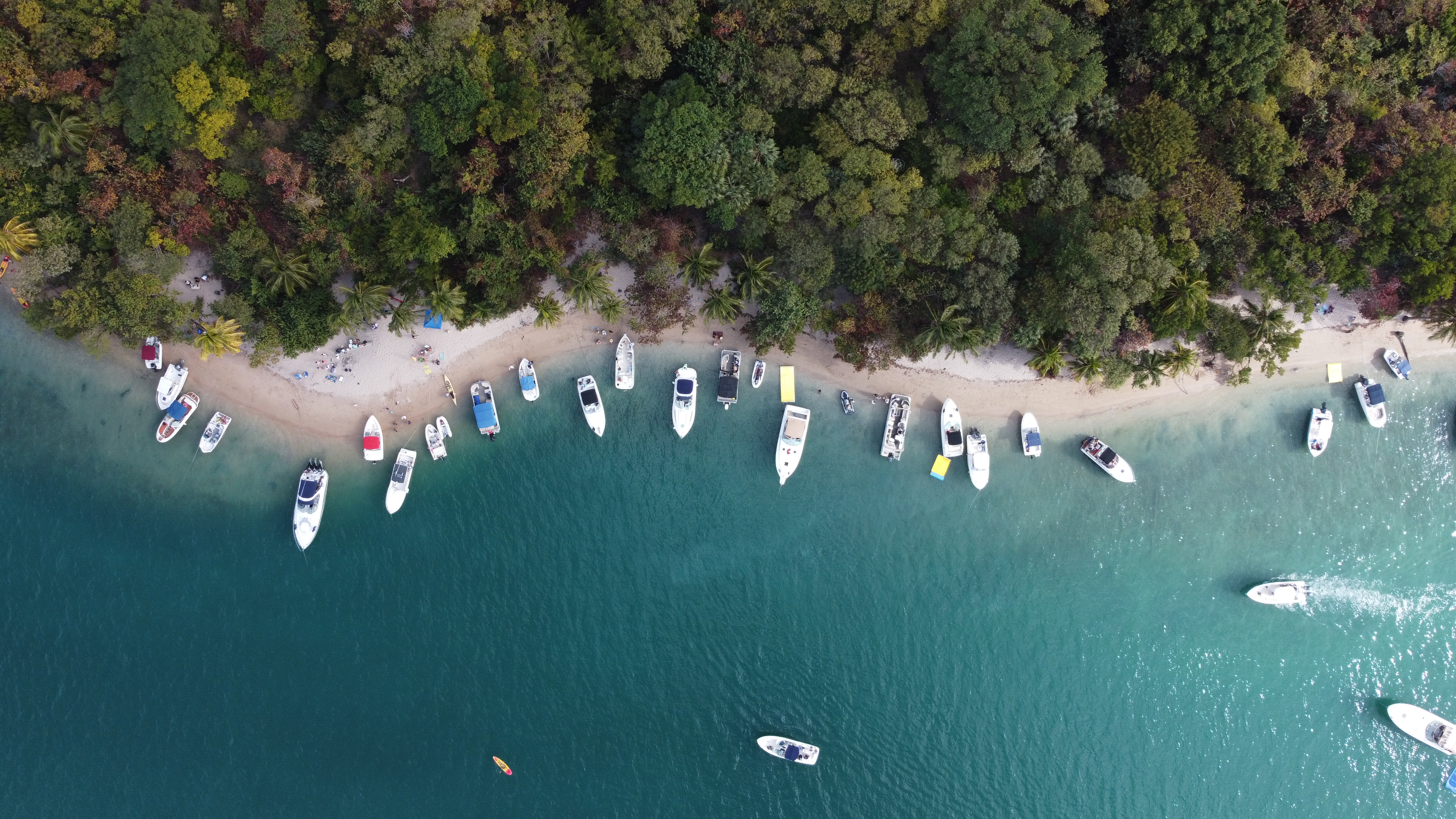 An aerial view of a tropical island with a white sand beach lined with boats of various sizes. Lush green trees cover the island, and the turquoise water sparkles in the sunlight. Picture taken by a drone.