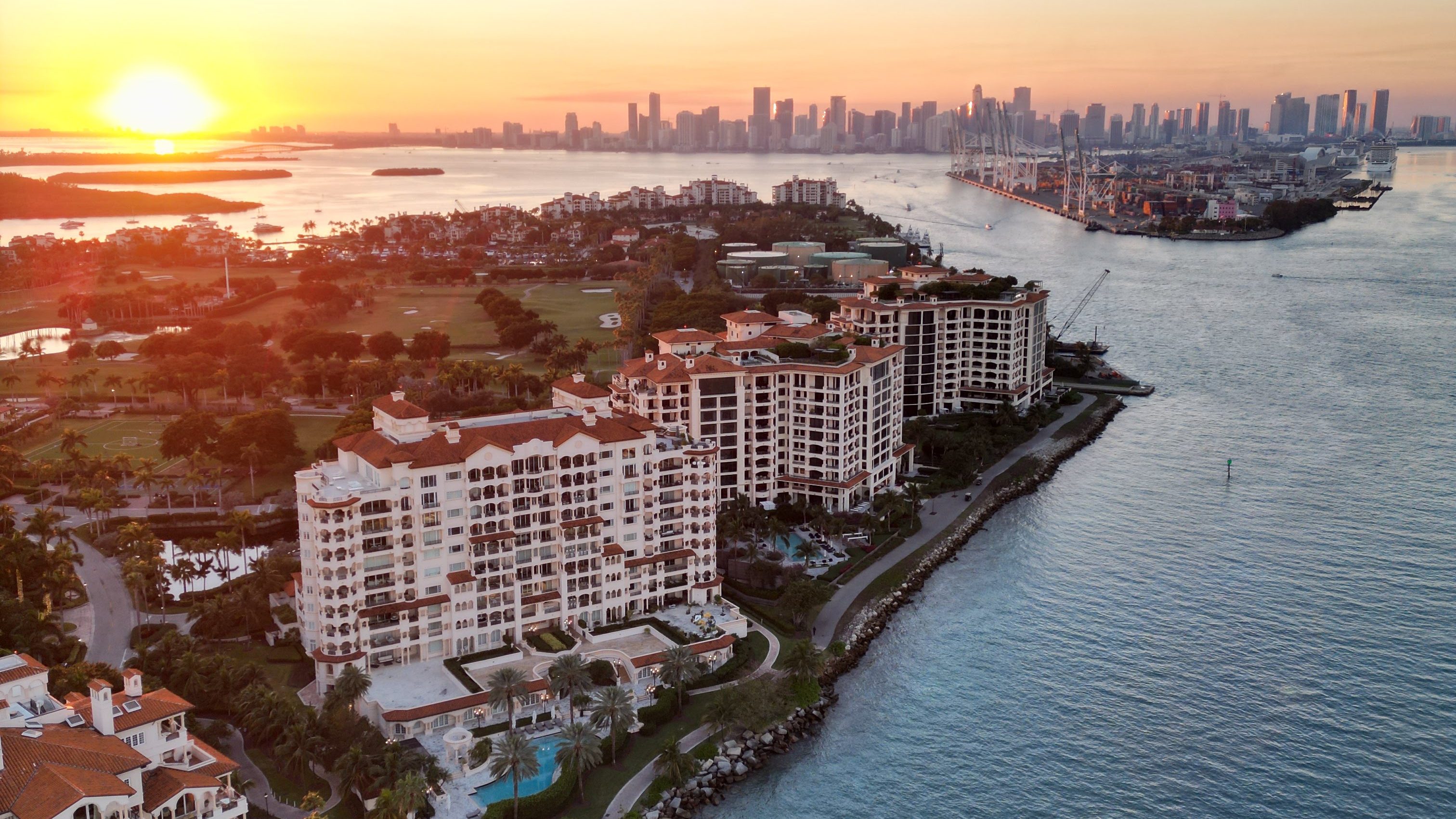 An aerial view of a luxury waterfront development with several high-rise buildings, swimming pools, and lush landscaping. The Miami skyline is visible in the distance. Picture captured by a drone.